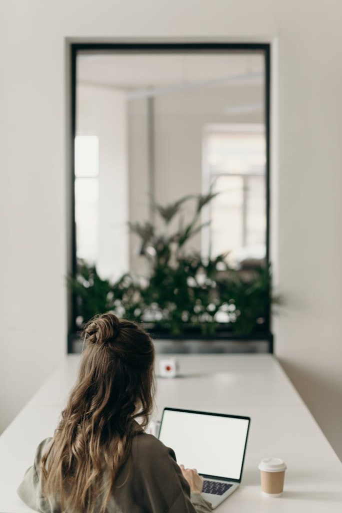 pexels photo 4065624 A woman with curly hair works on a laptop, facing a window with plants, symbolizing remote work.